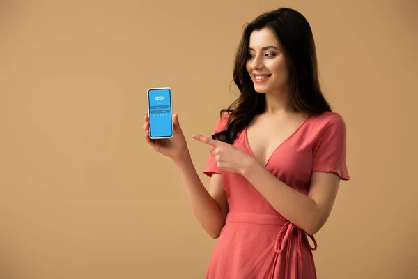 Alegre morena chica apuntando con el dedo en el teléfono inteligente con skype aplicación en la pantalla aislado en marrón - foto de stock