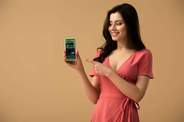 Menina morena alegre apontando com o dedo para o smartphone com aplicativo de reserva na tela isolada no marrom — Fotografia de Stock