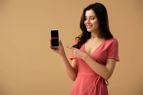 Весела блондинка вказуючи пальцем на смартфон з netflix app на екрані ізольовані на коричневий — Stock Photo