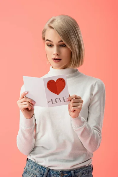 Surprised blonde woman looking at greeting card while standing isolated on pink — Stock Photo