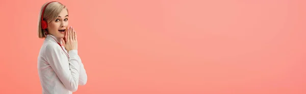 Панорамный снимок потрясенной блондинки, слушающей музыку в наушниках, изолированных на розовом — стоковое фото