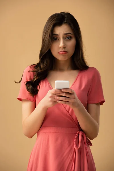 Chica morena molesta sosteniendo teléfono inteligente aislado en marrón - foto de stock