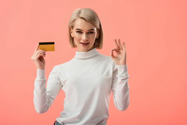 Щаслива блондинка молода жінка тримає кредитну картку і показує знак ОК ізольовано на рожевому — стокове фото