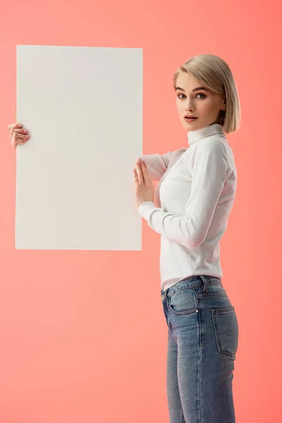 Femme blonde surprise tenant une pancarte blanche isolée sur rose — Photo de stock