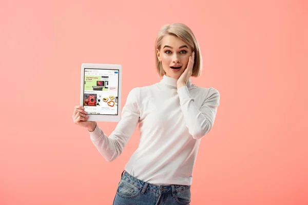 Mujer rubia sorprendida sosteniendo tableta digital con aplicación ebay en la pantalla aislada en rosa - foto de stock