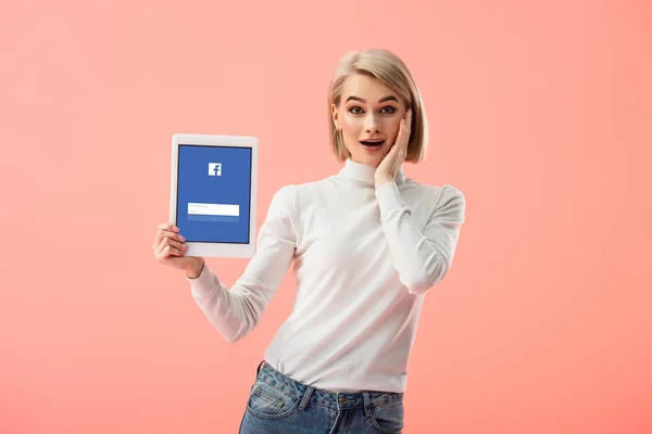 Здивований блондинка Жінка тримає Цифрова табличка з facebook app на екрані ізольовані на рожевий — стокове фото