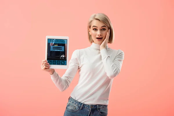 Здивована блондинка тримає цифровий планшет з додатком бронювання на екрані ізольовано на рожевому — стокове фото