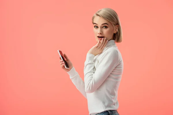 Здивована блондинка використовує смартфон ізольовано на рожевому — стокове фото