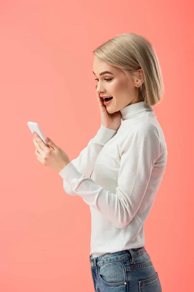 Shocked blonde girl looking at smartphone isolated on pink — Stock Photo