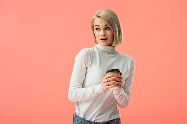 Impactada chica rubia sosteniendo taza de papel con bebida aislada en rosa - foto de stock