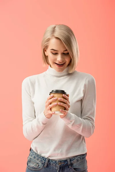 Chica rubia sorprendida mirando taza de papel con bebida aislada en rosa — Stock Photo