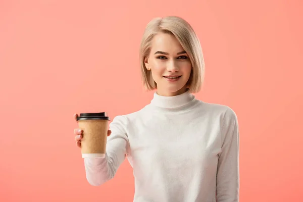 Enfoque selectivo de alegre chica rubia sosteniendo taza de papel con bebida aislada en rosa — Stock Photo