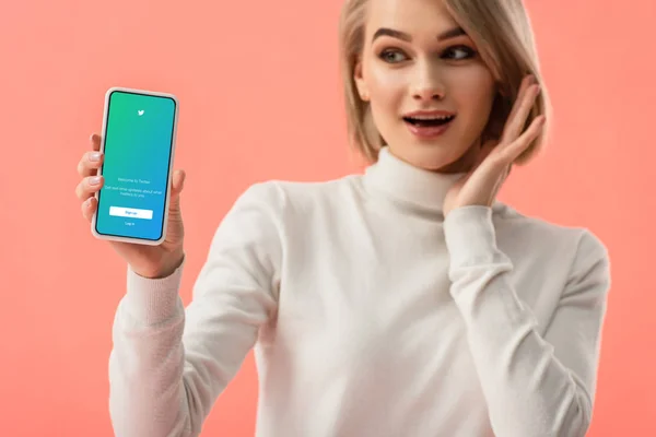 Selektiver Fokus einer überraschten blonden Frau, die ihr Smartphone mit Twitter-App auf dem Bildschirm isoliert auf rosa hält — Stockfoto
