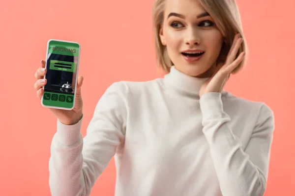Selektiver Fokus einer überraschten blonden Frau, die ihr Smartphone mit Buchungs-App auf dem Bildschirm isoliert auf pink hält — Stockfoto