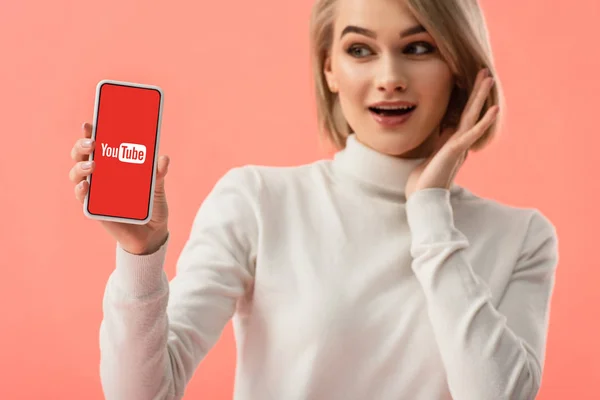 Foyer sélectif de femme blonde surprise tenant smartphone avec application youtube à l'écran isolé sur rose — Photo de stock