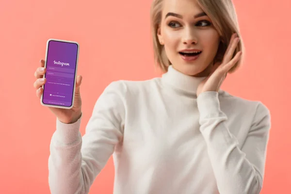 Foyer sélectif de femme blonde surprise tenant smartphone avec application instagram à l'écran isolé sur rose — Photo de stock