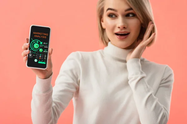 Enfoque selectivo de mujer rubia sorprendida sosteniendo smartphone con análisis de marketing en pantalla aislada en rosa - foto de stock