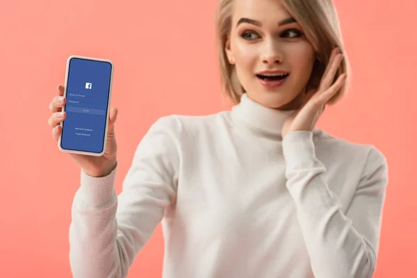 Messa a fuoco selettiva della donna bionda sorpresa che tiene lo smartphone con l'app facebook sullo schermo isolato su rosa — Foto stock