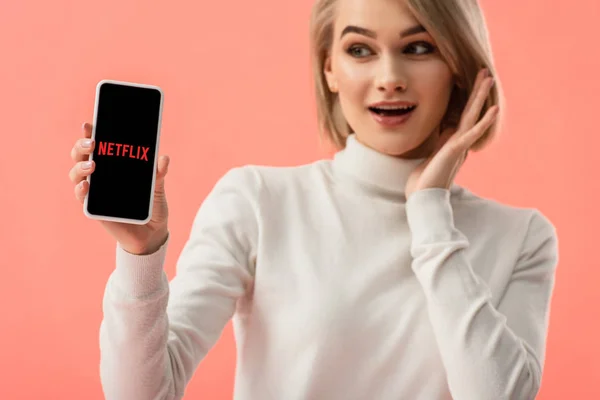 Селективный фокус удивленной блондинки, держащей смартфон с приложением netflix на экране изолирован на розовый — стоковое фото