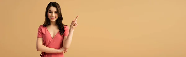 Panoramic shot of cheerful brunette woman pointing with finger while standing isolated on brown — Stock Photo