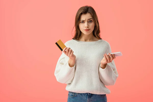 Confuso ragazza bruna in possesso di carta di credito e smartphone isolato su rosa — Foto stock