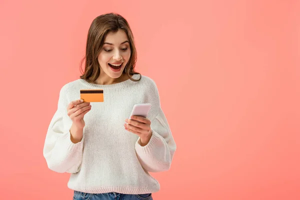 Überraschte Brünette hält Kreditkarte und Smartphone isoliert auf rosa — Stockfoto
