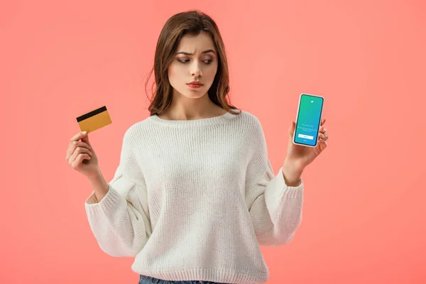 Menina morena atraente segurando cartão de crédito enquanto olha para o smartphone com aplicativo twitter na tela isolada no rosa — Fotografia de Stock