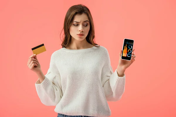 Menina morena atraente segurando cartão de crédito enquanto olha para o smartphone com gráficos e gráficos na tela isolada no rosa — Fotografia de Stock