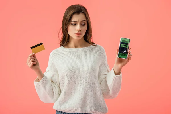 Atractiva chica morena sosteniendo tarjeta de crédito mientras mira el teléfono inteligente con aplicación de reserva en la pantalla aislada en rosa - foto de stock