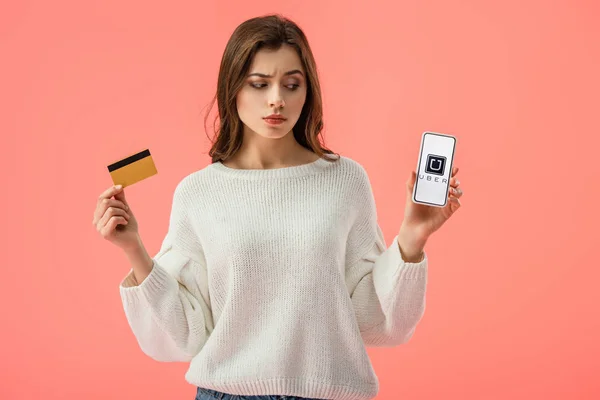 Atractiva chica morena sosteniendo la tarjeta de crédito mientras mira el teléfono inteligente con la aplicación uber en la pantalla aislada en rosa - foto de stock