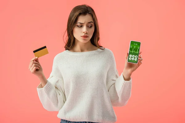 Menina morena atraente segurando cartão de crédito enquanto olha para o smartphone com aplicativo de saúde na tela isolada no rosa — Fotografia de Stock