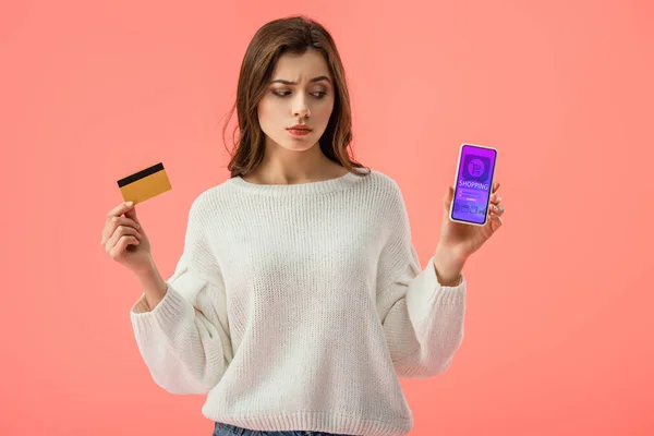 Приваблива брюнетка дівчина тримає кредитну картку, дивлячись на смартфон із додатком для покупок на екрані ізольовано на рожевому — стокове фото