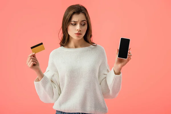 Attractive brunette girl holding credit card while looking at smartphone with blank screen isolated on pink — Stock Photo