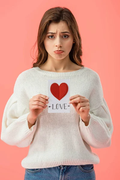 Triste joven mujer celebración tarjeta con amor letras aislado en rosa - foto de stock