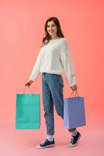 Vista completa de la mujer en suéter blanco y jeans sosteniendo bolsas de compras sobre fondo rosa - foto de stock