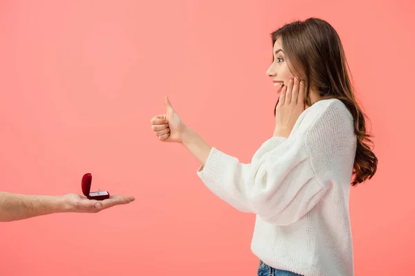 Vista recortada del hombre haciendo propuesta de matrimonio a chica morena sorprendida con el pulgar hacia arriba aislado en rosa - foto de stock