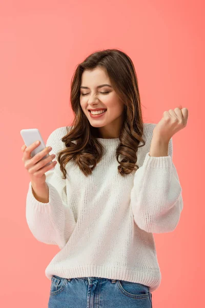 Mujer feliz y atractiva en suéter blanco sosteniendo teléfono inteligente aislado en rosa - foto de stock