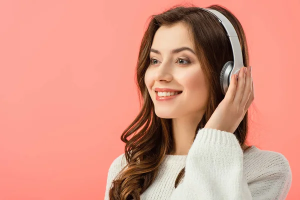 Mujer sonriente y hermosa en suéter blanco escuchando música con auriculares aislados en rosa - foto de stock