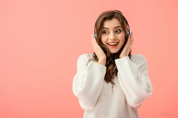 Smiling and beautiful woman in white sweater listening music with headphones isolated on pink — Stock Photo