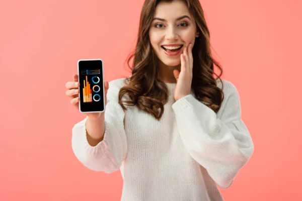 Enfoque selectivo de la mujer en suéter blanco que sostiene el teléfono inteligente con gráficos y gráficos en la pantalla aislada en rosa - foto de stock