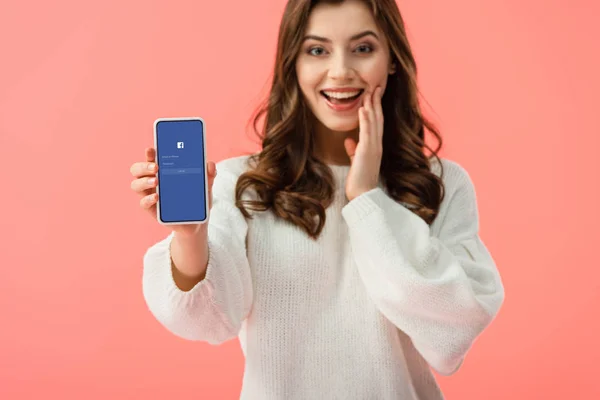Селективное внимание женщины в белом свитере, держащей смартфон с приложением facebook на экране, изолированном на розовый — стоковое фото