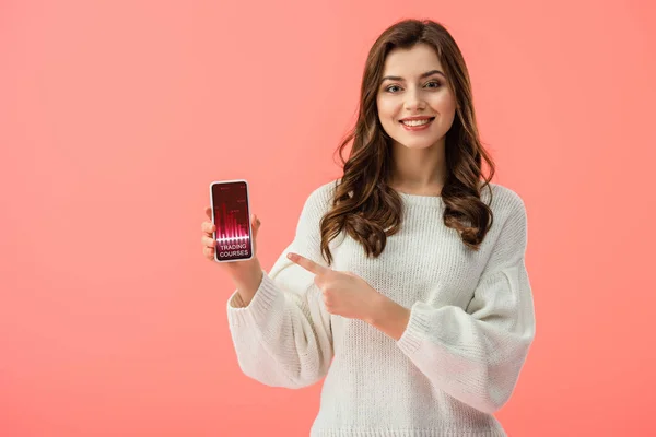 Mulher em camisola branca apontando com o dedo para o smartphone com cursos de negociação aplicativo na tela isolada no rosa — Fotografia de Stock