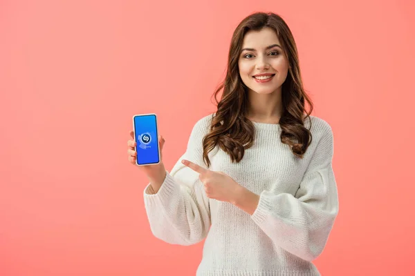 Frau im weißen Pullover zeigt mit dem Finger auf Smartphone mit Shazam-App auf Bildschirm isoliert auf rosa — Stockfoto