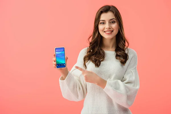 Woman in white sweater pointing with finger at smartphone with booking app on screen isolated on pink — Stock Photo