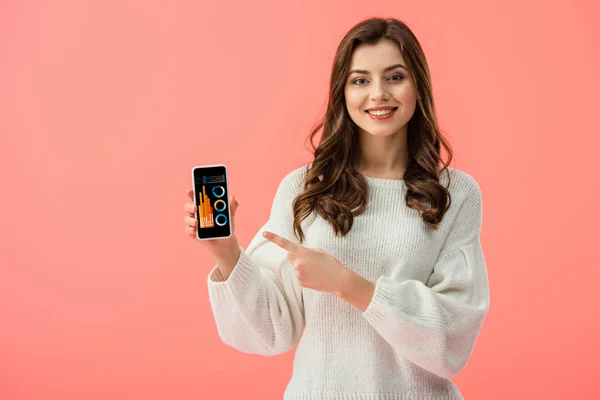 Woman in white sweater pointing with finger at smartphone with charts and graphs on screen isolated on pink — Stock Photo