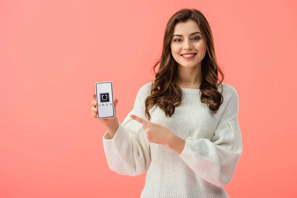 Mujer en suéter blanco apuntando con el dedo al teléfono inteligente con aplicación uber en la pantalla aislada en rosa - foto de stock