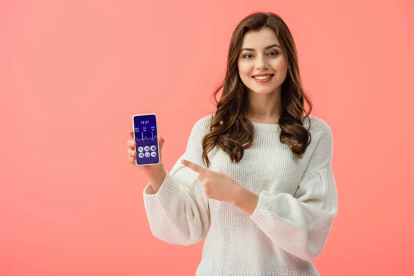 Mujer en suéter blanco apuntando con el dedo al teléfono inteligente con aplicación de salud en la pantalla aislada en rosa - foto de stock