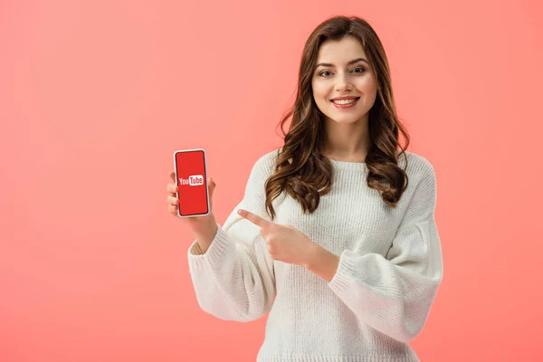 Mujer en suéter blanco apuntando con el dedo al teléfono inteligente con aplicación de youtube en la pantalla aislada en rosa - foto de stock