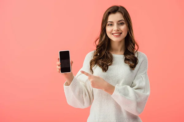 Mujer en suéter blanco apuntando con el dedo al teléfono inteligente con pantalla en blanco aislado en rosa - foto de stock