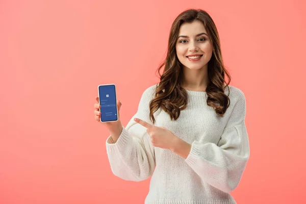 Mujer en suéter blanco apuntando con el dedo al teléfono inteligente con aplicación de Facebook en la pantalla aislada en rosa - foto de stock